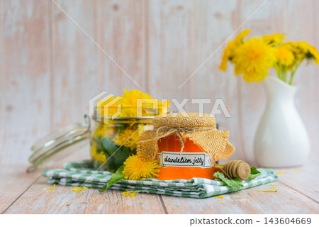 Transparent glass of jar DANDELION JELLY text with dandelion flowers in vase. Soft natural lighting highlights the amber color glossy texture and artisanal character of the preserve. Concept of Transparent glass of jar DANDELION JELLY text with dandelion flowers in vase. Soft natural lighting highlights the amber color glossy texture and artisanal character of the preserve. Concept of 143604669