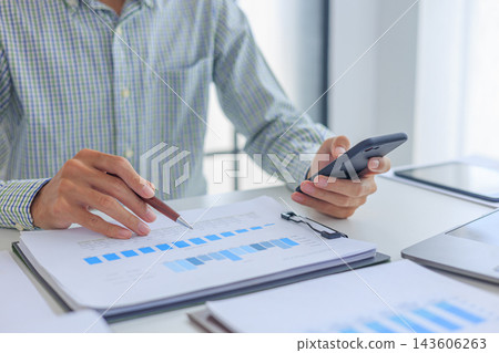 Businessman working in office using smartphone while reviewing and analyzing graph data at desk. Businessman working in office using smartphone while reviewing and analyzing graph data at desk. 143606263