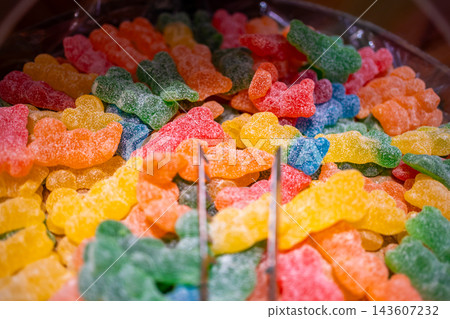 Metal tongs pick up a yellow sugar coated gummy bear from a container of colorful candies. Pick and mix confectionery selection in a candy shop with selective focus Metal tongs pick up a yellow sugar coated gummy bear from a container of colorful candies. Pick and mix confectionery selection in a candy shop with selective focus 143607232