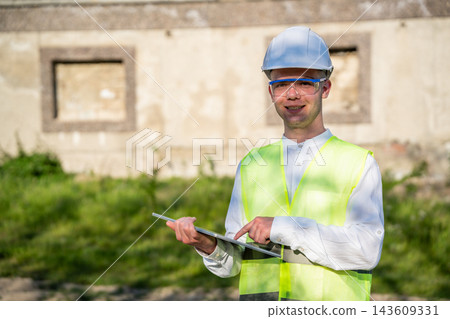 Construction worker in safety vest and hard hat using tablet at building site. Site operations digital construction documents and mobile project management 143609331