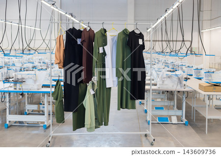 Clothes hanging on a rack in a clothing factory with sewing machines in the background Clothes hanging on a rack in a clothing factory with sewing machines in the background 143609736