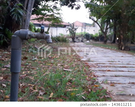 Old metal spigot mounted on pvc pipe. Static water faucet in backyard. Outdoor landscape design with greenery. Dry autumn leaves on ground. Old metal spigot mounted on pvc pipe. Static water faucet in backyard. Outdoor landscape design with greenery. Dry autumn leaves on ground. 143611211