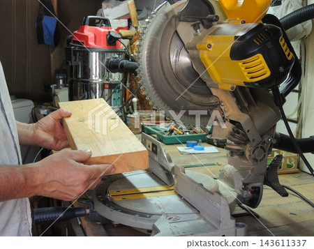 The craftsman holds a thick wooden block, positioning it against the bed of a miter saw to make a precise cut. A professional construction vacuum cleaner is visible in the background. 143611337