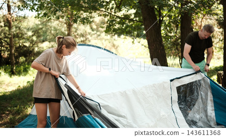 Father and daughter are pitching a tent in a wooded area during daytime 143611368