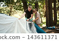 Father and daughter work together to assemble a tent in a forested setting under sunlight 143611371