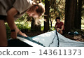 Mother and daughter set up a tent in a forest clearing surrounded by trees 143611373