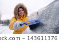 Woman in a yellow jacket removes snow using a brush on a cold day in a parking lot 143611376