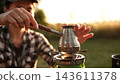 Woman Making Coffee In A Cezve On A Portable Camping Stove Outdoors At Sunset 143611378
