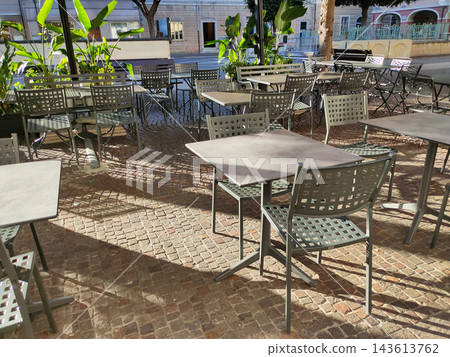 Outdoor cafe terrace with empty metal tables and chairs on a rustic cobblestone pavement, surrounded by tropical plants and historic European buildings in warm sunlight. Outdoor cafe terrace with empty metal tables and chairs on a rustic cobblestone pavement, surrounded by tropical plants and historic European buildings in warm sunlight. 143613762