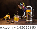 Dark mood tea still life with illuminated double wall glass cup, tea drops frozen in air, french press, dry flowers, ginger root, and spices on dark wooden table. 143613863