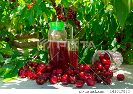 Jug of cherry juice and ripe cherries on table under tree leaves Jug of cherry juice and ripe cherries on table under tree leaves 143617953