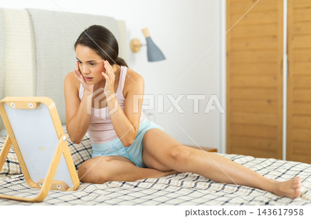 Attentive young girl looking at her face in mirror in bedroom Attentive young girl looking at her face in mirror in bedroom 143617958