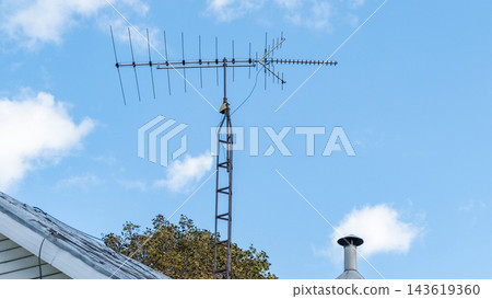 Rooftop television antenna rising against a bright blue sky with clouds beside a chimney and weathered roof Rooftop television antenna rising against a bright blue sky with clouds beside a chimney and weathered roof 143619360