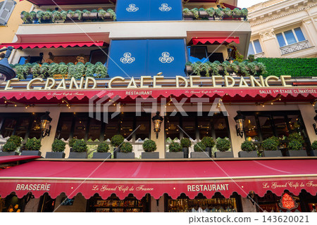 Street level view of Le Grand Cafe de France in Nice, France, with deep red awnings, navy balcony panels, string lights, and layered terraces in early evening. Street level view of Le Grand Cafe de France in Nice, France, with deep red awnings, navy balcony panels, string lights, and layered terraces in early evening. 143620021