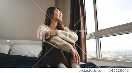 Happy young adult asian woman relax sitting on bed, wearing knitwear looking out window 143620042
