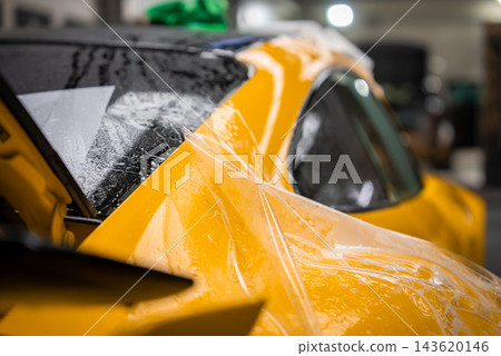 Close view of a yellow luxury sports car as clear film is applied on a wet soapy panel, showing water beads, squeegee marks, and a lifted edge under soft indoor light. Close view of a yellow luxury sports car as clear film is applied on a wet soapy panel, showing water beads, squeegee marks, and a lifted edge under soft indoor light. 143620146
