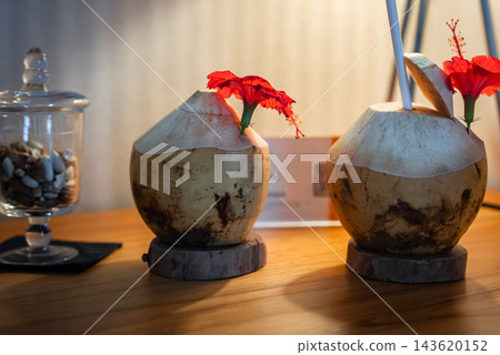 Two coconuts with hibiscus flowers and a straw sit on coasters on a polished table at a Maldives resort, with a jar of nuts on the left and warm ambient lighting. Two coconuts with hibiscus flowers and a straw sit on coasters on a polished table at a Maldives resort, with a jar of nuts on the left and warm ambient lighting. 143620152