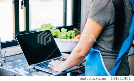 A woman stands in the kitchen happily cooking while using a laptop for video tutorials 143621461