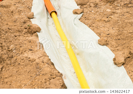 A trench with protective fabric is ready for a drainage pipe installation. Gravel and soil are strategically placed around the pipe for efficient water management. A trench with protective fabric is ready for a drainage pipe installation. Gravel and soil are strategically placed around the pipe for efficient water management. 143622417