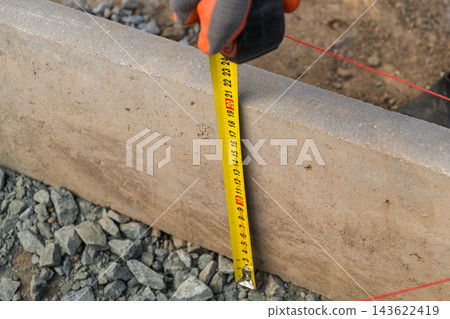 A tape measure stretches out as it is held in a hand, indicating the precise length of a concrete block on a gravel surface at a construction site. A tape measure stretches out as it is held in a hand, indicating the precise length of a concrete block on a gravel surface at a construction site. 143622419