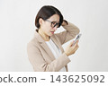 A middle-aged businesswoman with her smartphone in her hands, holding her head in her hands, against a white background. 143625092