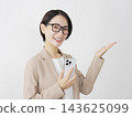 A middle-aged businesswoman holding a smartphone and pointing to the right with her palm against a white background. 143625099