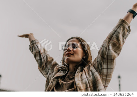 Woman raining happiness raising arms to the sky while standing outdoors during light rain shower under grey sky 143625264