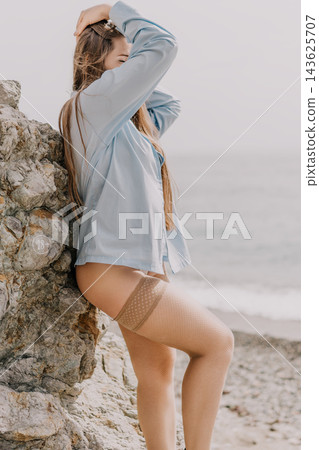 Woman beach stockings leaning against a coastal rock while wearing a blue shirt and fishnet hosiery by the ocean Woman beach stockings leaning against a coastal rock while wearing a blue shirt and fishnet hosiery by the ocean 143625707