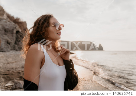 Woman Beach Sunset Woman in sunglasses and white swimsuit stands on a rocky beach at sunset enjoying serene vacation vibes 143625741