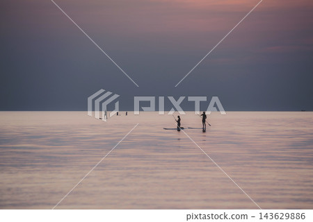 夕陽西下，紫色的海水映襯著人們在立式槳板上衝浪的剪影。 143629886