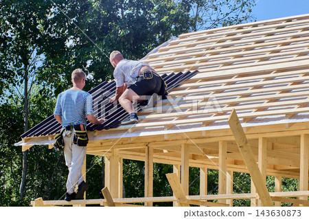 Asphalt roofing sheets are installed by two construction workers who position bitumen panels across wooden roof rafters during house construction. Asphalt roofing sheets are installed by two construction workers who position bitumen panels across wooden roof rafters during house construction. 143630873