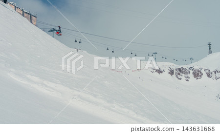 Gondola lift crossing snowy mountain slope with skiers and rocky ridgeline under pale winter sky 143631668