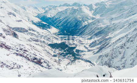 Panoramic snowy alpine valley with ski resort village nestled between towering snow-covered peaks in bright winter daylight and mo Panoramic snowy alpine valley with ski resort village nestled between towering snow-covered peaks in bright winter daylight and mo 143631754