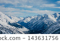 Panoramic view of snow-covered Mount Elbrus and surrounding Caucasus peaks under pale blue sky, deep glacial valley leading toward 143631756