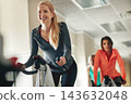 Woman, aerobics bike and coach at gym, training and support for fitness in cycling class. Female person, riding and stationary machine for workout challenge, spin instructor and equipment at club 143632048