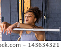 African American female leaning on Olympic barbell rack, wearing grey tanktop and smartwatch in gym 143634321