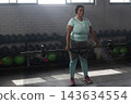 African American female lifting loaded Olympic barbell across thighs in gym with dumbbell rack 143634554