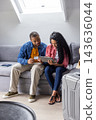 Couple sitting on light-gray sofa beneath skylight, holding tablet and using card near suitcase 143636044