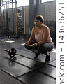 African American woman crouching on mat in gym near loaded barbell wearing tanktop fitness watch 143636251