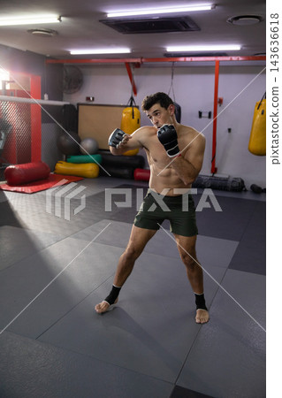 Practicing shirtless fighter wearing green shorts, wraps striking in MMA gym with cage and bags 143636618
