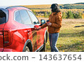 Woman opening red car door holding coffee cup during autumn road trip. Traveler stopped in countryside with forest landscape background 143637654