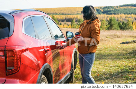Woman opening red car door holding coffee cup during autumn road trip. Traveler stopped in countryside with forest landscape background Woman opening red car door holding coffee cup during autumn road trip. Traveler stopped in countryside with forest landscape background 143637654