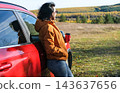 Woman standing near red car holding coffee cup during autumn road trip, resting and enjoying scenic autumn landscape. Travel and freedom concept 143637656