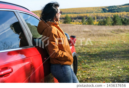 Woman standing near red car holding coffee cup during autumn road trip, resting and enjoying scenic autumn landscape. Travel and freedom concept 143637656