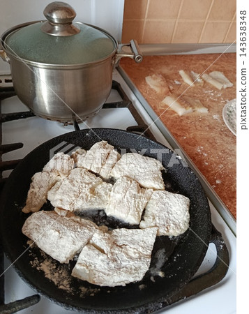 Fish fillets coated in flour cooking in a hot cast iron frying pan on a gas stove burner next to a lidded pot, preparing a delicious seafood meal 143638348
