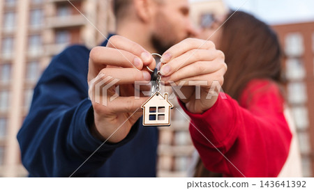 Closeup Couple Kissing And Holding Keys With House Keychain Outside Apartment Building. First Home Purchase, Mortgage, Homeownership And Shared Achievement Concept. Closeup Couple Kissing And Holding Keys With House Keychain Outside Apartment Building. First Home Purchase, Mortgage, Homeownership And Shared Achievement Concept. 143641392