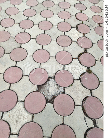 Outdoor paved walkway featuring a geometric tessellation of circular pink-red and star-patterned grey concrete tiles. The aged surface shows wear, cracks, and weeds in the joints. Outdoor paved walkway featuring a geometric tessellation of circular pink-red and star-patterned grey concrete tiles. The aged surface shows wear, cracks, and weeds in the joints. 143642634