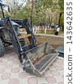 Detailed close-up of a blue and black front loader with its bucket resting on circular paving stones in an urban park. Hydraulic pistons and background trees are visible. 143642635