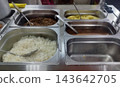 Buffet line with stainless steel trays. Contents include rice, mashed potatoes, meat sauce, and buckwheat. Several serving spoons are placed inside the trays. 143642705