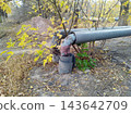 Weathered metal utility pipes with damaged insulation elbows run along the ground in a neglected, autumnal outdoor setting near trees. 143642709